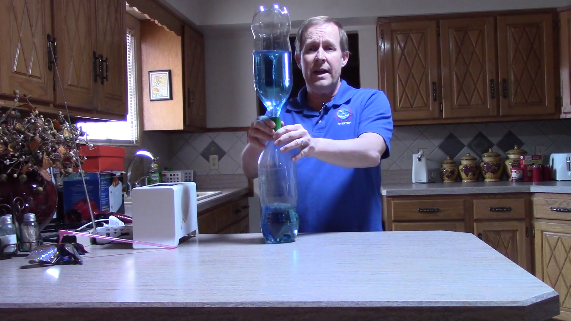  A person wearing a National Weather Service shirt and standing in a home kitchen holds two plastic 1-liter bottles that are stacked on top of each other vertically, connected at the neck, and partially filled with colored liquid. A funnel shape, like that of a tornado, forms in the liquid in the upper bottle as it flows through the narrow neck and into the lower bottle. 