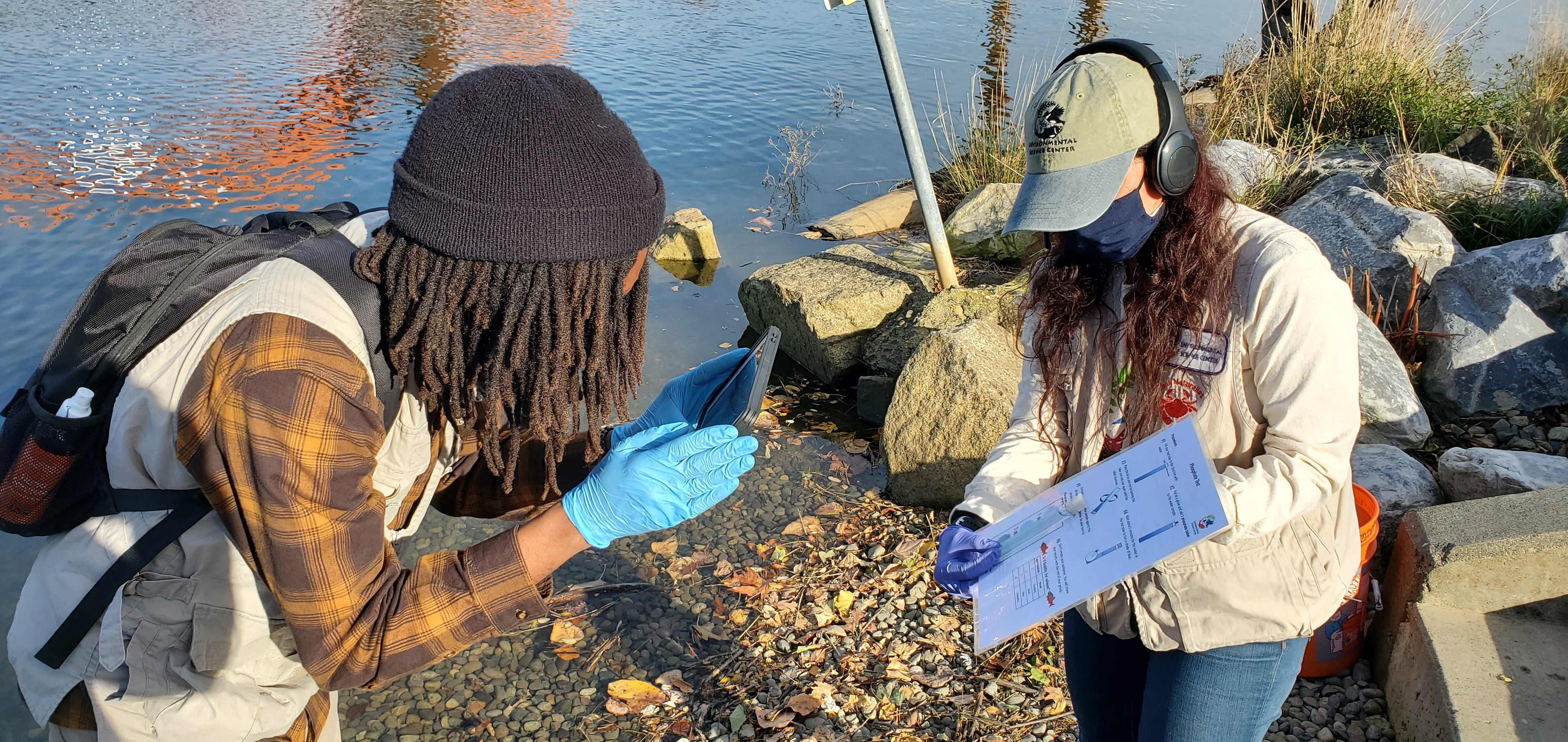 Two people stand along a stream. One is holding a data sheet and a tube of liquid held up next to the sheet to compare colors. The second person is filming the other with a tablet. Both wear protective masks and gloves.