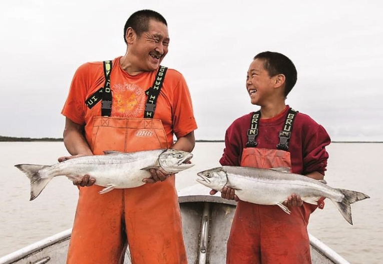 An indigenous man and boy in orange fishing gear are holding fish and smiling at each other.