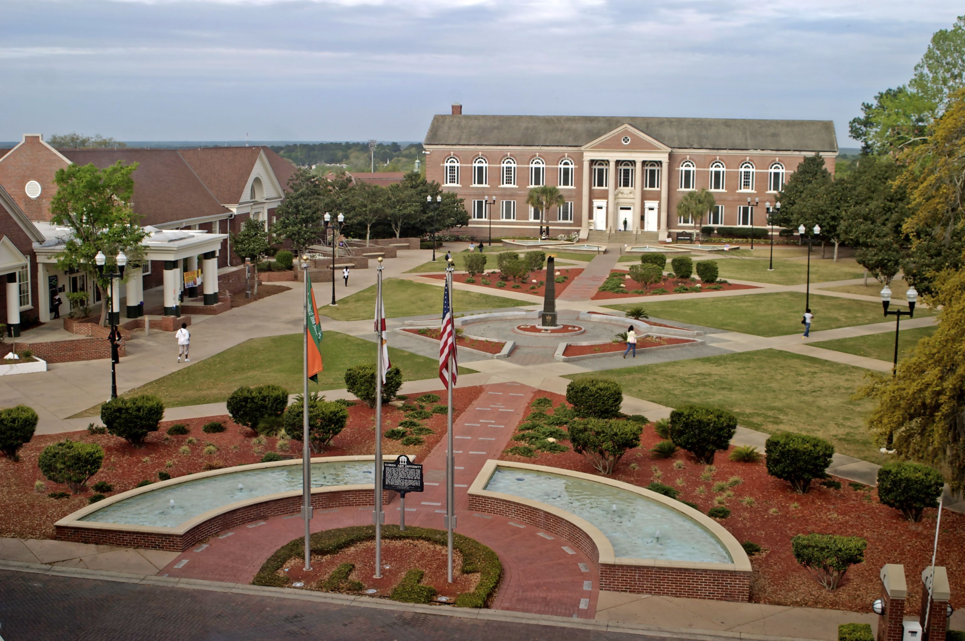 A photo of a college campus that looks down on the campus as if the photo was taken from an upper story. The campus has central lawn with decorative geometric walkways with modern fountains at the entrance. Buildings surround the central lawn.