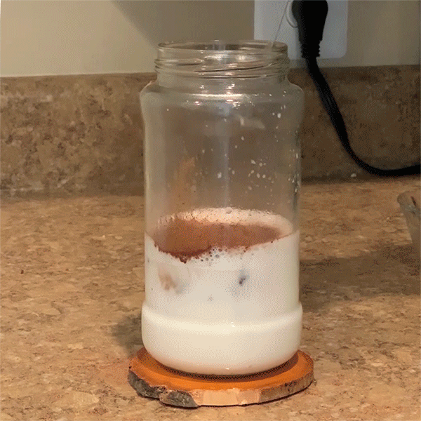 A looping GIF showing a close-up view of hot chocolate in a mason jar being mixed by a handheld milk frother. As the milk froths, pale foam builds up on top of the hot chocolate.