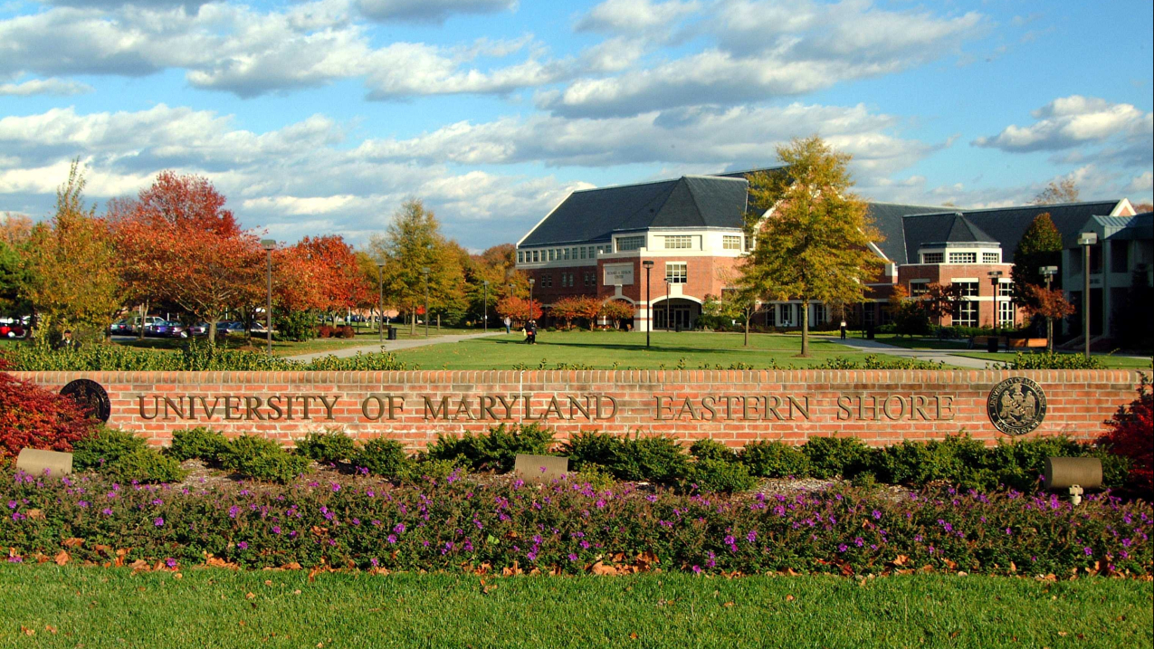 A short brick wall with lettering that reads "University of Maryland Eastern Shore." Behind it, there is a nicely landscaped lawn leading to a college campus. 