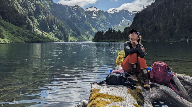 Emma Rudy sits on a smooth weathered log that is lying in shallow water and smiles, looking up at the sky. The water flows into a mountainous Alaskan landscape behind her.