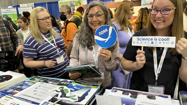 Two smiling teachers hold up signs, one with the NOAA logo, the other reads "Science is cool!," while standing in front of a table covered with educational materials. Other teachers are milling about and visiting other booths behind them.