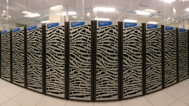 Image is of the NOAA Hera supercomputer located in Fairmont, WV. Hera has 52,640 Skylake processors with multiple grey cabinets (20 are shown in the image) and is labeled with CRAY CS500. The racks of hardware components are stacked vertically to save space,  allow for ease of connecting the many nodes and cores of the supercomputer, and improve efficiency in cooling.