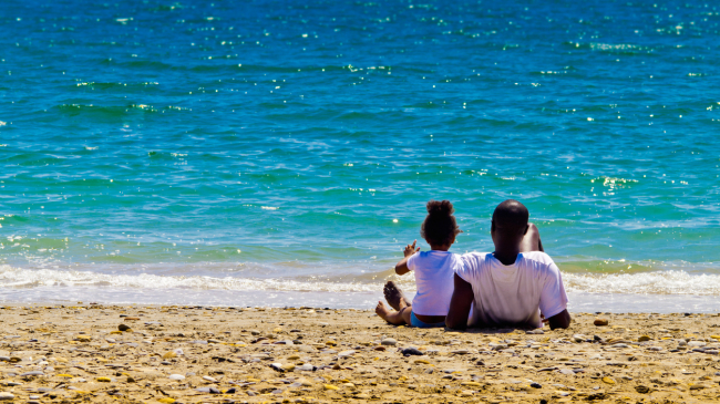 A father and his young daughter wearing white t-shirts and bathing suits enjoy a quiet moment alone sitting on a stretch of beach with turquoise waters on a very bright and sunny day. (stock image.)