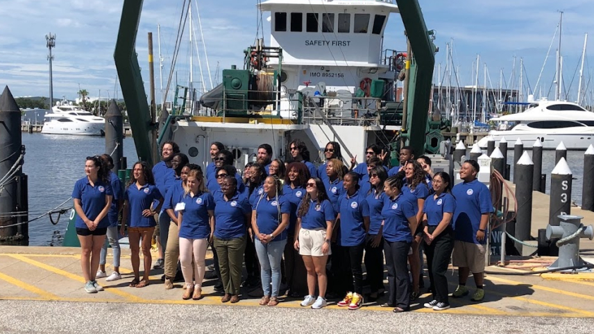 A group of people in matching blue shirts with CCME logos pose in front of a large, docked vessel.