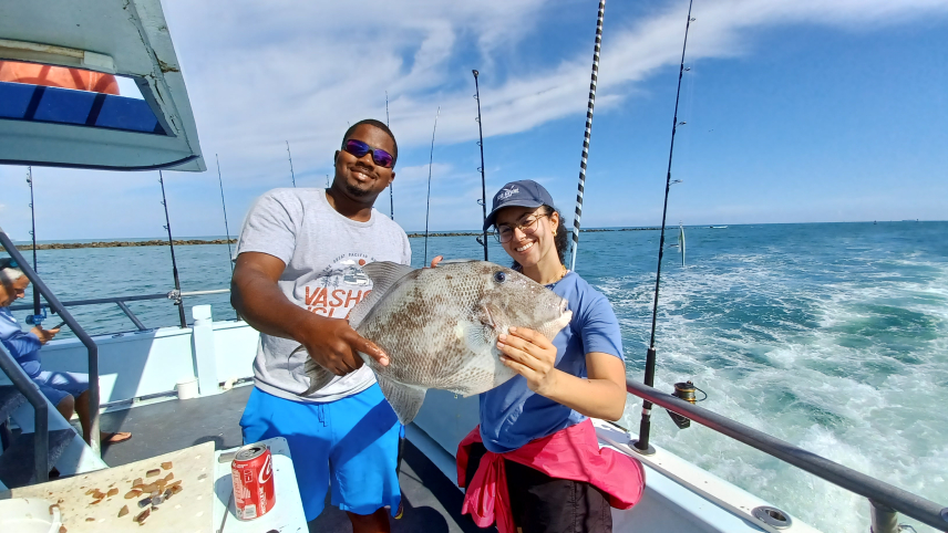 Two people on a fishing boat hold a triggerfish, one person holds the tail and the other it's abdomen. 