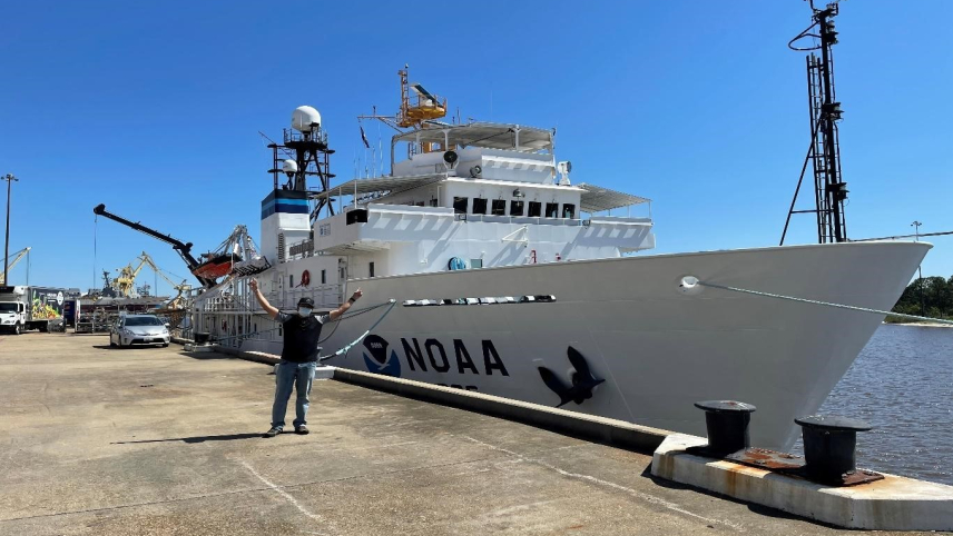  Eder Herrera, wearing a mask, stands on a dock in front of a large NOAA research vessel. He appears small compared to the size of the ship, and his hands are raised and reaching out enthusiastically. 