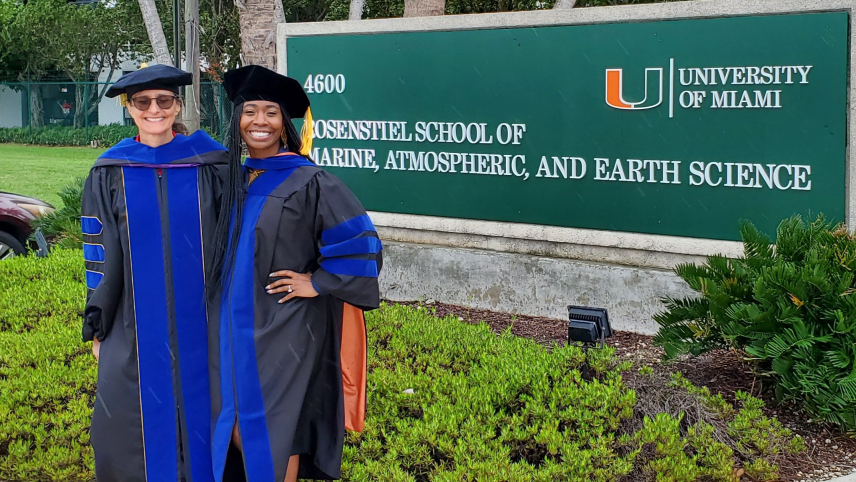 Adrianne Wilson and Elizabeth Babcock, both wearing academic regalia, stand with their arms around each other in front of a sign for the University of Miami Rosenstiel School of Marine, Atmospheric, and Earth Sciences. 