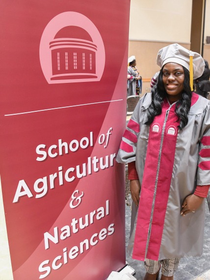 Jasmine Smalls wearing academic regalia stands by a banner that says, "School of Agriculture & Natural Sciences" in an auditorium that appears to be set up for a graduation ceremony.