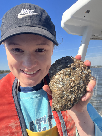 Kaitlynn Wade smiles while holding an oyster up for the camera. She is on a boat in bay and wearing a baseball hat and a floatation device. 