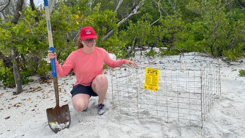 Helen poses with her hand on a cage that will cover a sea turtle nesting area. In her other hand, she holds a shovel.