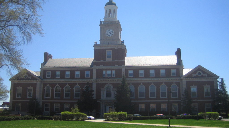 A three-story brick building with a clock tower, which is topped in a dome that rises to a spike.