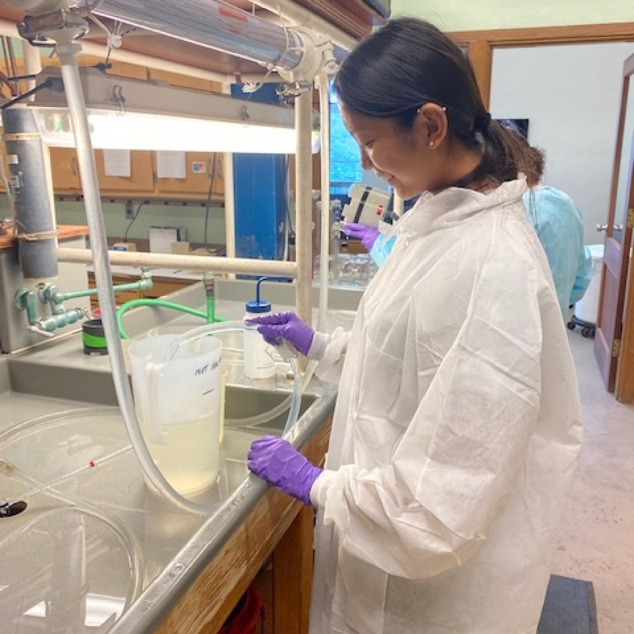 Rose wears a white lab coat and rubber gloves and is in what looks like a chemistry lab. She is pouring liquid into a pitcher through a tube.