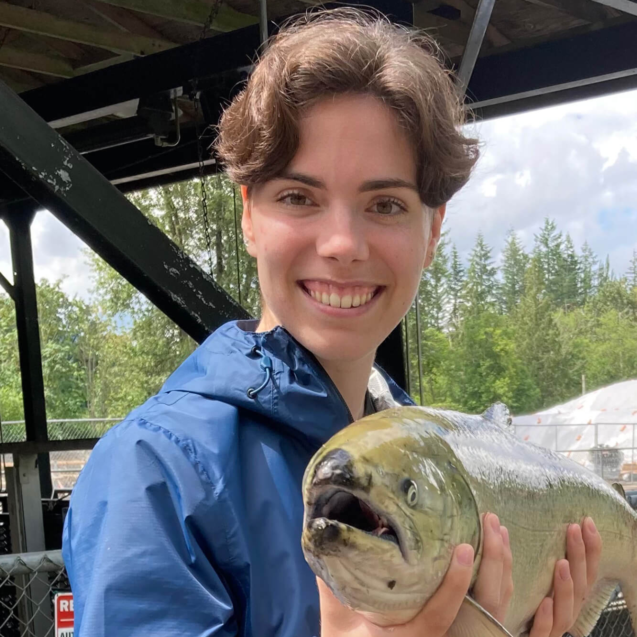 Magdalena smiles at the camera and holds a salmon so that it is looking into the camera.