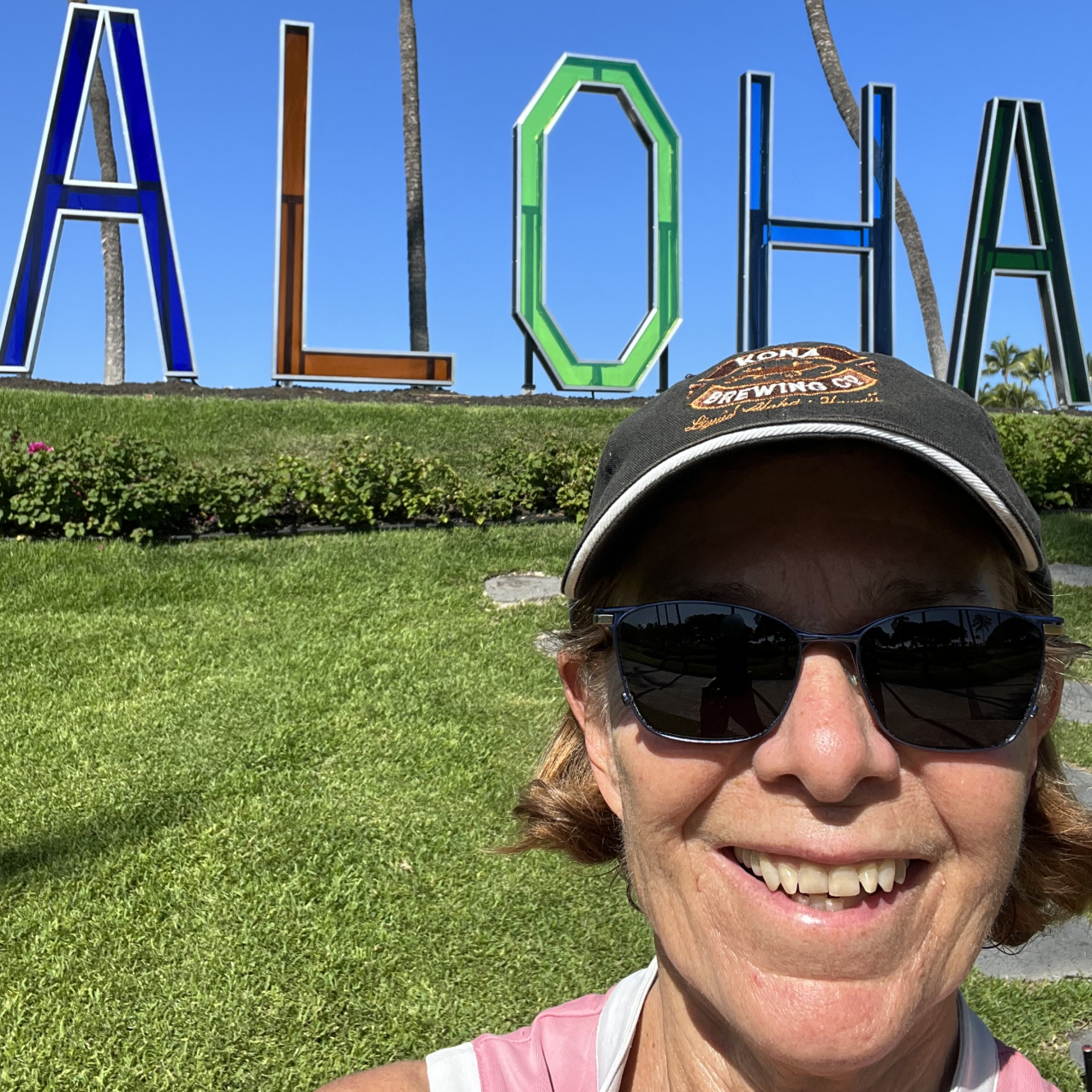 Mary-Sue Atkinson, wearing a hat and sunglasses, stands in front of a large outdoor sign that says "Aloha." 