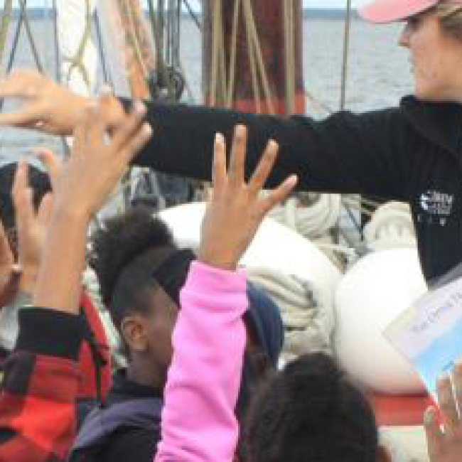 Students are on a boat in a river raising their hands as an educator talks to them.