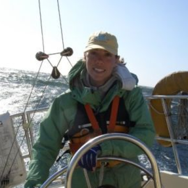 Amber stands at the wheel of a vessel at sea wearing cold weather field clothes and smiling at the camera.