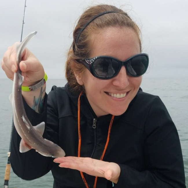 Cara holds up a small shark that looks to be about a foot long and smiles. She is standing next to a fishing pole on an overcast day.