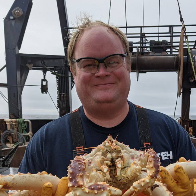 Kevin stands on a vessel at sea. He is wearing waders and thick rubber gloves, and he is holding a large crab that looks to be a few feet long.