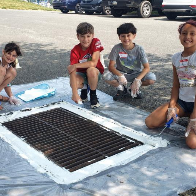 four students sit around a drain on the street with cars in the background. The students are wearing gloves and holding paint brushes. Around the drain is fresh white paint.