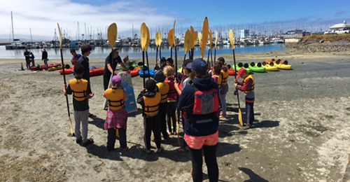 A group of children stand on a beach all holding kayak paddles and wearing yellow life jackets.