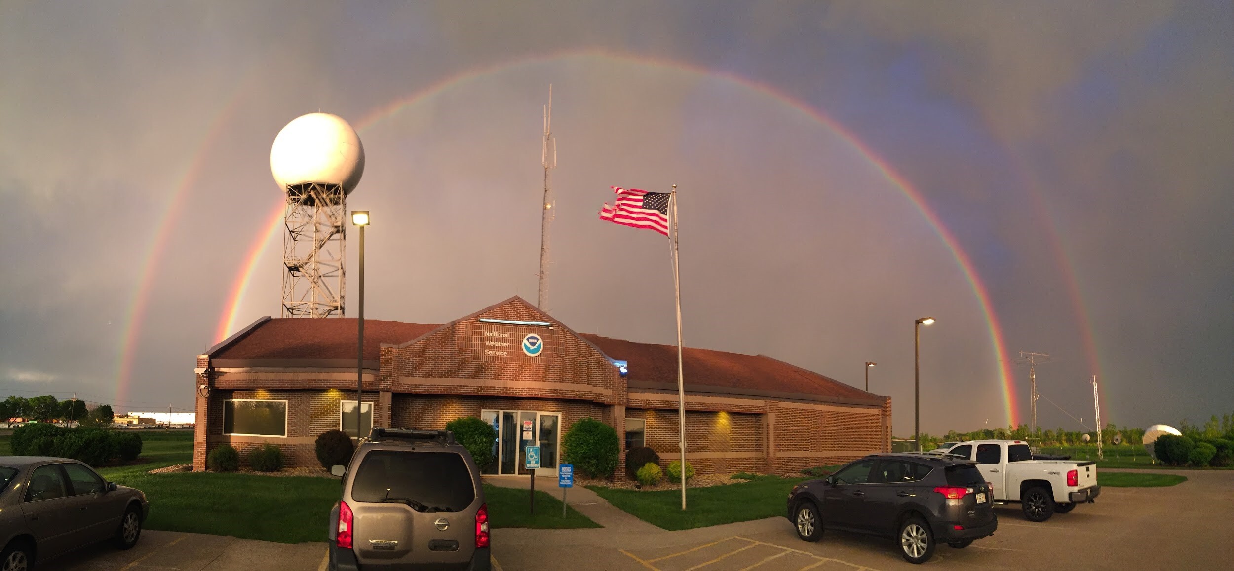  A double rainbow forms over a building with the NOAA and National Weather Service logos, an American flag, and a weather radar. 