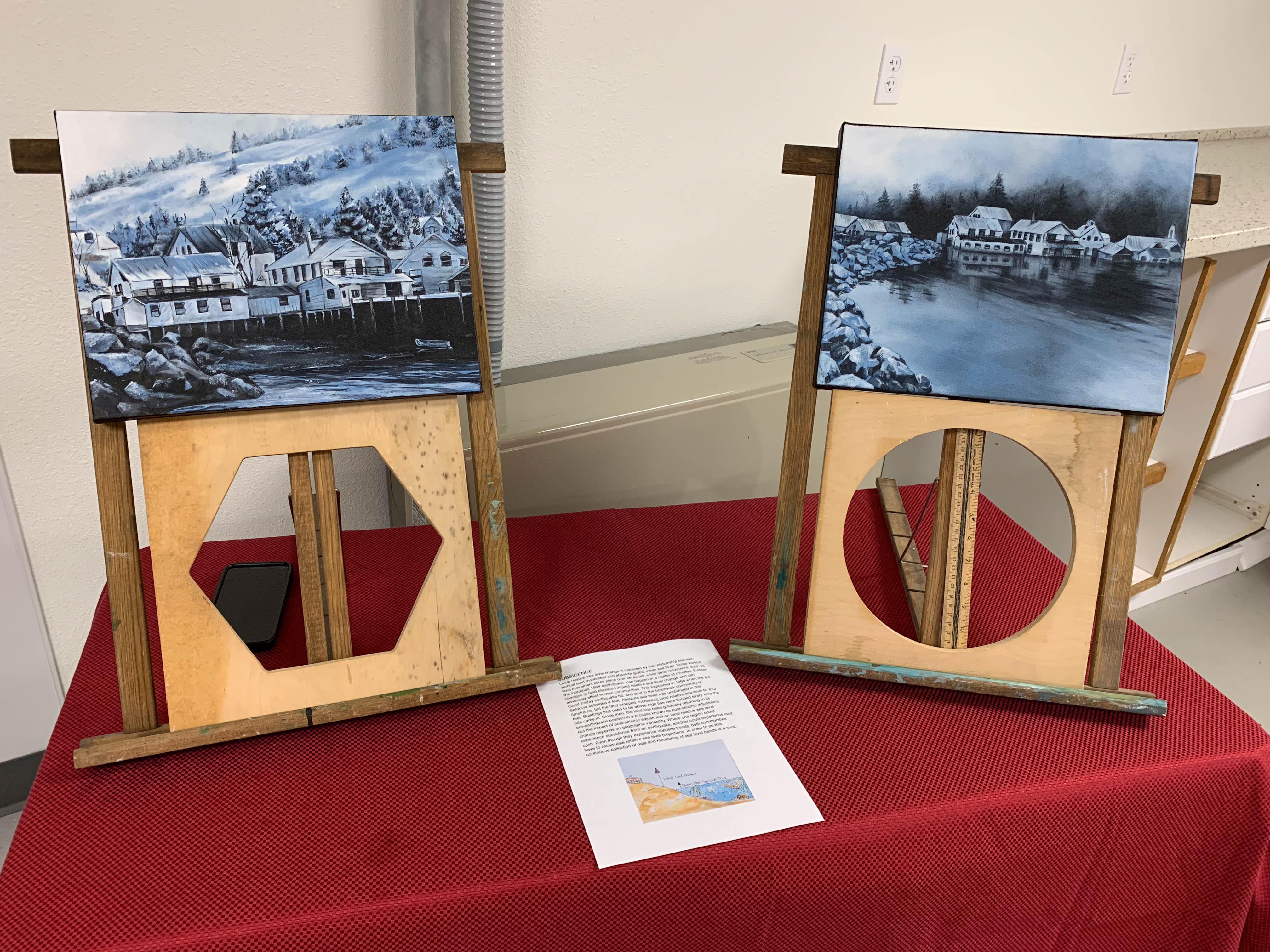Two paintings displayed on a table. On the left is a snowy coastal town with a mountainous landscape behind it.  The painting rests on a wooden frame with a cut out hexagon. The right painting shows the same town from a more distant location, but the coastal waters now flood the town by what appears to be a few feet above the elevated frames that many houses rested on. The painting is on a wooden frame with a circle cutout and a ruler in the circular opening. 