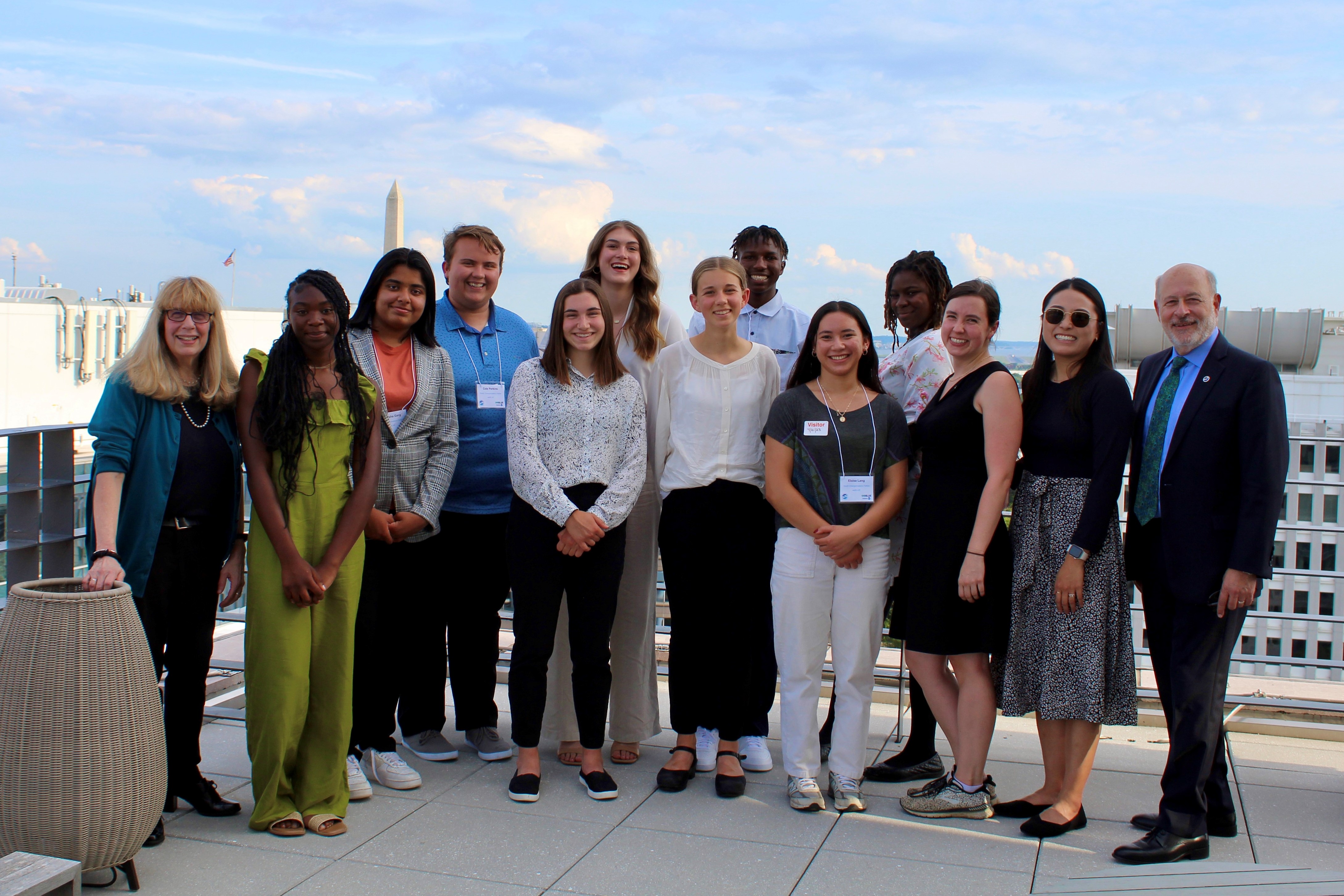 A group of 13 people, nine high school students and four adults, smile at the camera. They are standing on a rooftop patio in Washington, DC, with the Washington Monument in the background.