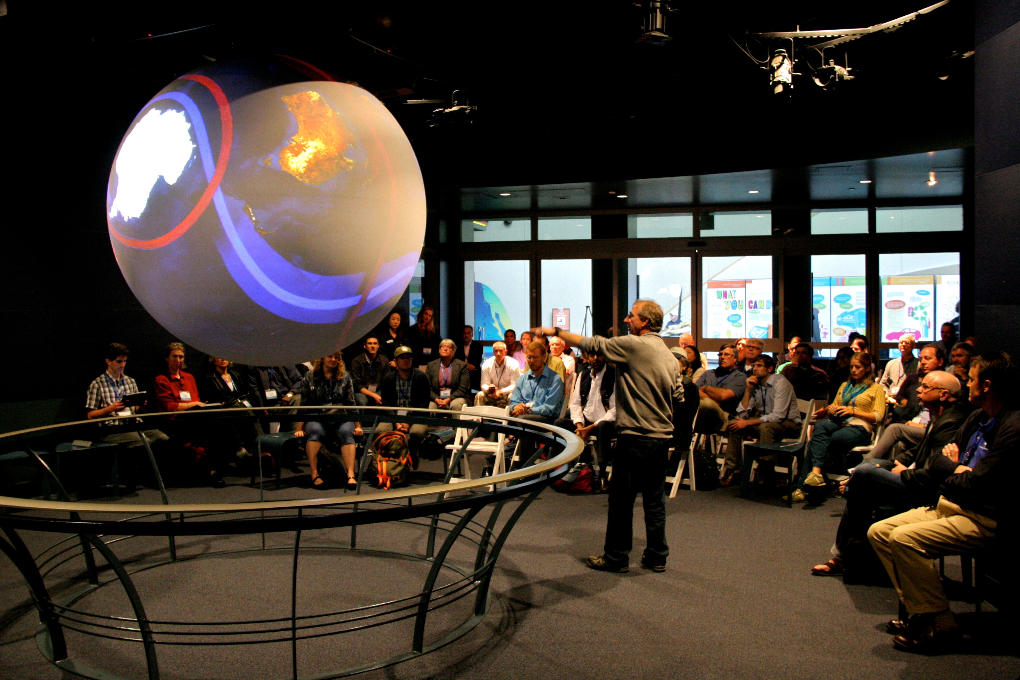 A presenter speaking to a group of people, gestoring towards a 6 foot sphere explaining earth science data.