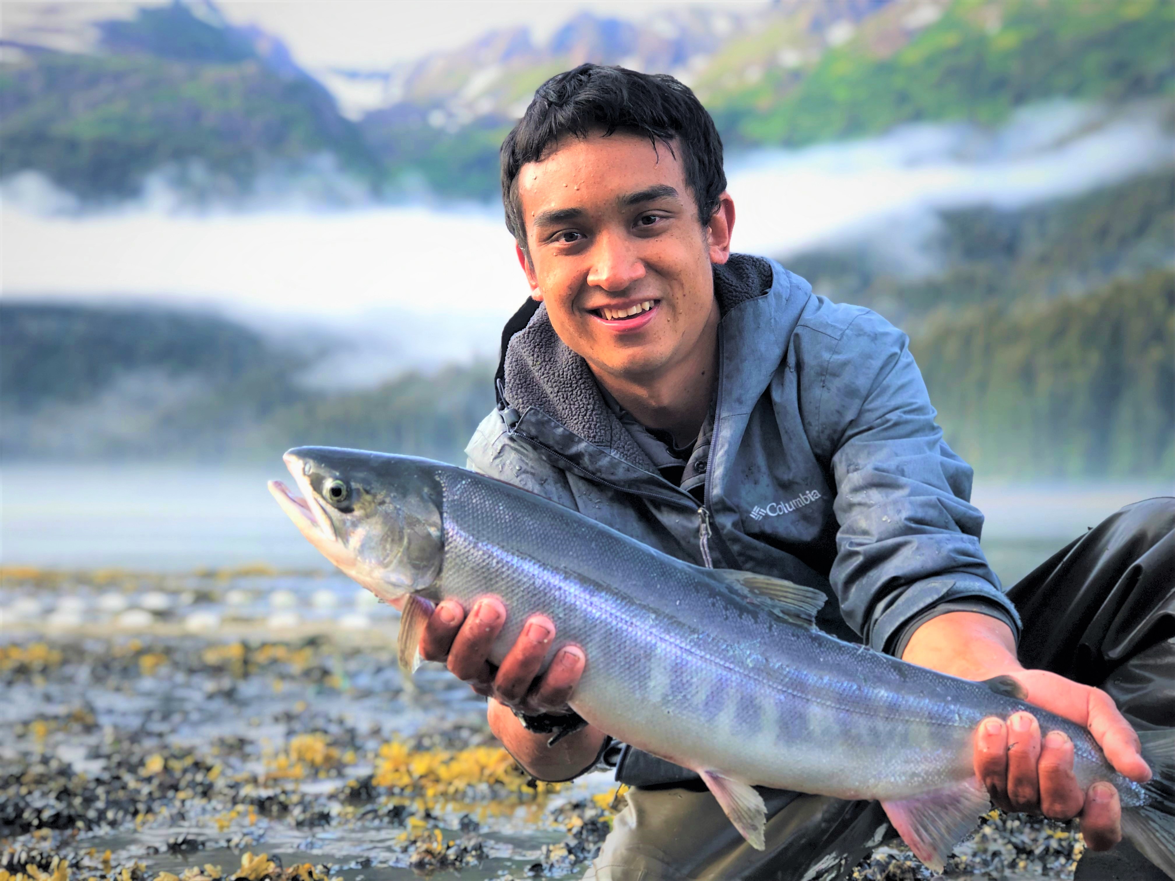 Andrew Tokuda, Global environmental science major at University of Hawaiʻi at Mānoa and 2018 NOAA Hollings scholar, holds a chum salmon while kneeling beside the Tutka Bay estuary. Tokuda interned at the Kachemak Bay National Estuarine Research Reserve in Homer, Alaska, in summer 2019.