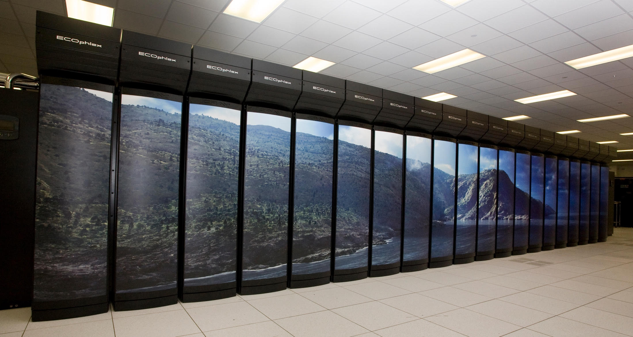 Image is of the NOAA Gaea supercomputer located in Oak Ridge, TN. Gaea is a supercomputer with 143,936 Intel cores and consists of multiple black cabinets (19 are shown in the image). The racks of hardware components are stacked vertically to save space, allow for ease of connecting the many nodes and cores of the supercomputer, and to improve efficiency in cooling. In this picture, an artwork is placed in front of the black cabinets showing picturesque hilly coastal scene which has been divided up on the