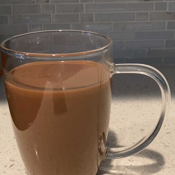 A looping GIF of milk being poured slowly from a measuring cup into a glass mug that is partially filled with cocoa. The milk is poured first into a spoon that is resting on the surface of the cocoa. As the mug fills up, a layer of pale milk forms on top of the darker cocoa.