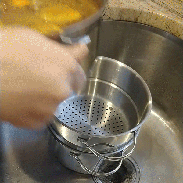 A looping GIF of cider containing orange slices and an assortment of whole spices being poured through a colander. The holes in the colander are about a quarter inch in diameter. The oranges and larger spices are caught in the colander while the cider drains through. 