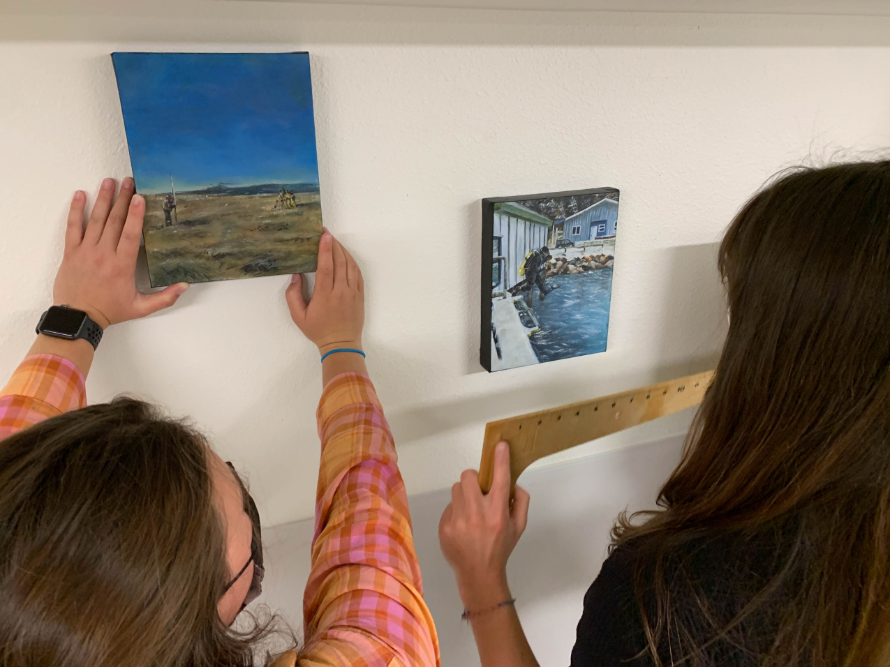 Kate holds a ruler as her sister places a painting. The left painting shows someone holding a pole that extends several feet above them while another person looks through a surveying tripod in a coastal area. The second painting shows a scuba diver mid-jump from a boat docked near the shore.