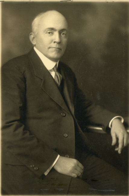 Black and white photo portrait of Nicholas H. Heck. He sits with his left arm resting on the arm of a chair, looking straight into the camera.