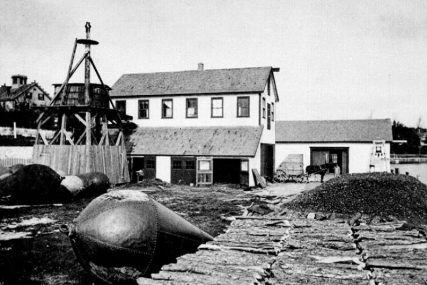 A black and white photo of the original Woods Hole, MA lab building with renovations, 1875. A crate housing a piano is visible on the horse-drawn carriage at right.
