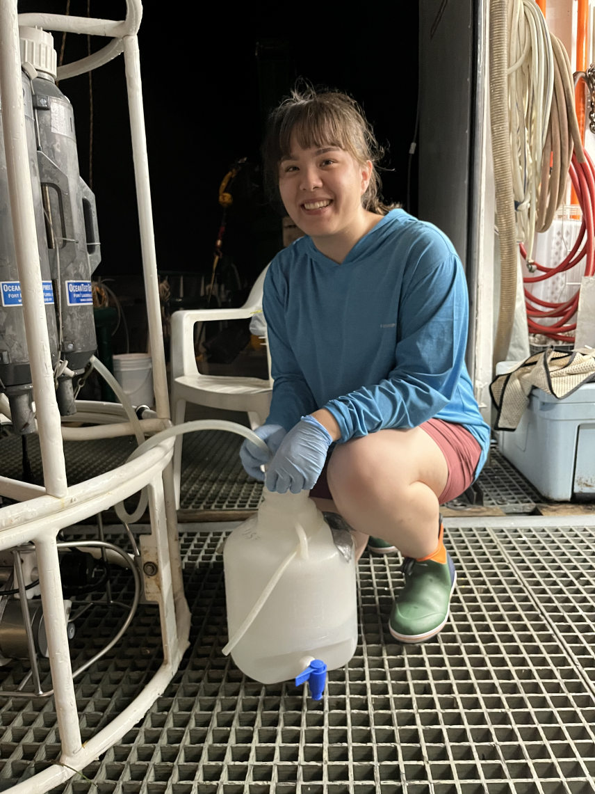 Anna smiles and kneels on the grated vessel floor. In a rubber gloved hand, Anna holds a tube coming from a large sampling device into the opening of a plastic jug with a spigot at the bottom.