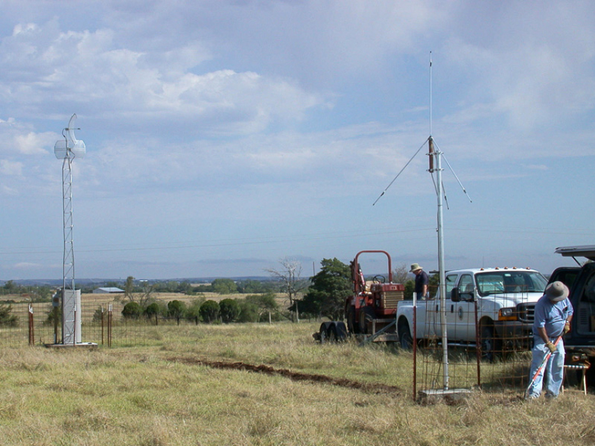 A photo of the Oklahoma Lightning Mapping Array (OKLMA) site north of Chickasha, OK. There is a VHF antenna on the right that sands signals to a small plastic building on the left, which houses electronics that process those signals.