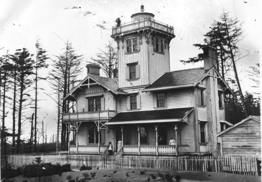 Black and white photo of the old Point Adams lighthouse, a wooden house with a tower in the middle that has the light on top of it. The lighthouse stands on the sand with scraggly trees behind it.