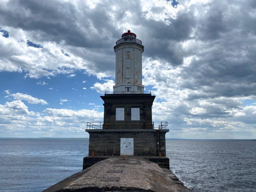 Photo of Keweenaw Waterway Lower Entrance Light in Chassell, MI. The lighthouse stands at the end of a pier, framed by water on either side. The tower is white and the foundation is red brick.