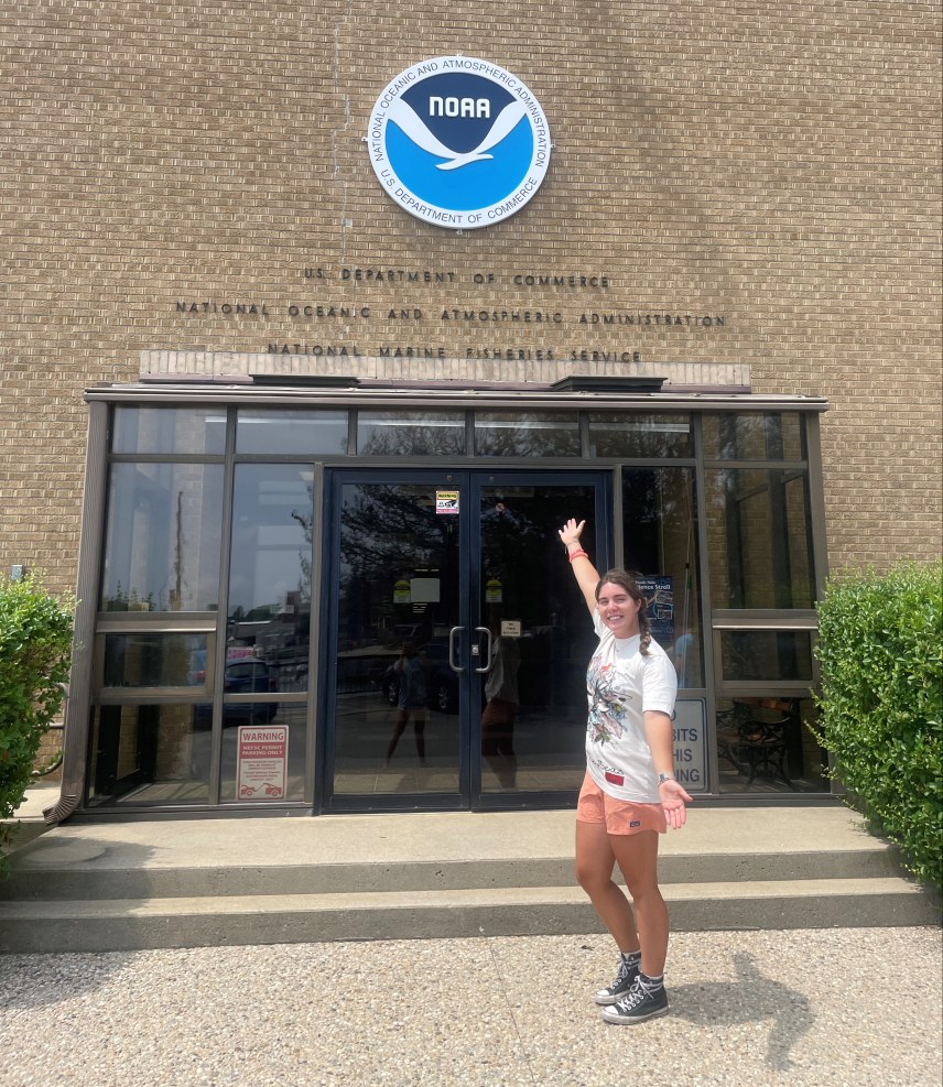 Cassidy poses, smiling with arms spread, in front of tan brick building with a large NOAA logo above the words "U.S. Department of Commerce, National Oceanic and Atmospheric Administration, National Marine Fisheries Service."