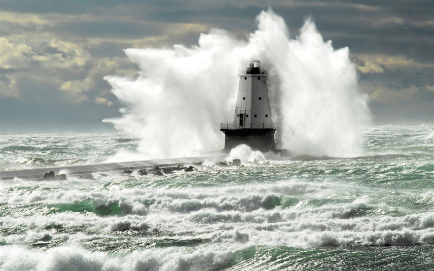 Photo of Ludington Lighthouse in Michigan. The white lighthouse sits in the ocean at the end of a pier. Waves taller than the lighthouse are crashing onto it.