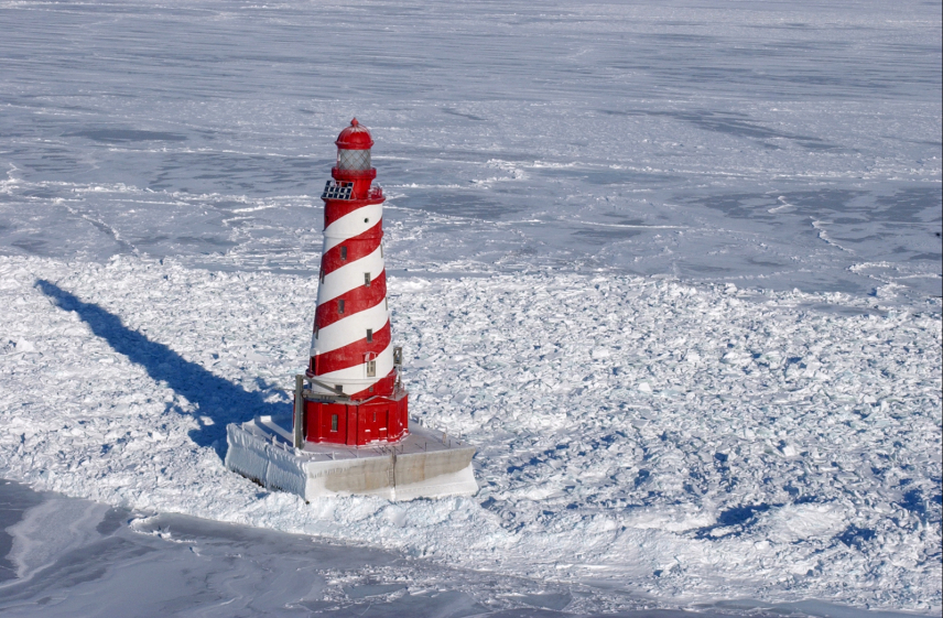 Photo of the White Shoal Light in Lake Michigan in the depths of winter. The lighthouse is candy-striped in red and white and sits on a square concrete base surrounded by snow.
