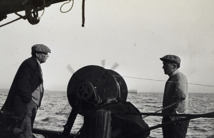 Black and white photo of Nicholas H. Heck in the field, as he performs  a wire-drag survey aboard the contract sloop Nena Rowland in 1913. He stands to the right of a large pulley, speaking with another man standing to the left.