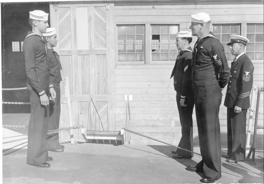 Black and white photo of a Chief Boatswain (far right) and crew members in formation as "Sideboys" to welcome a VIP aboard ship.