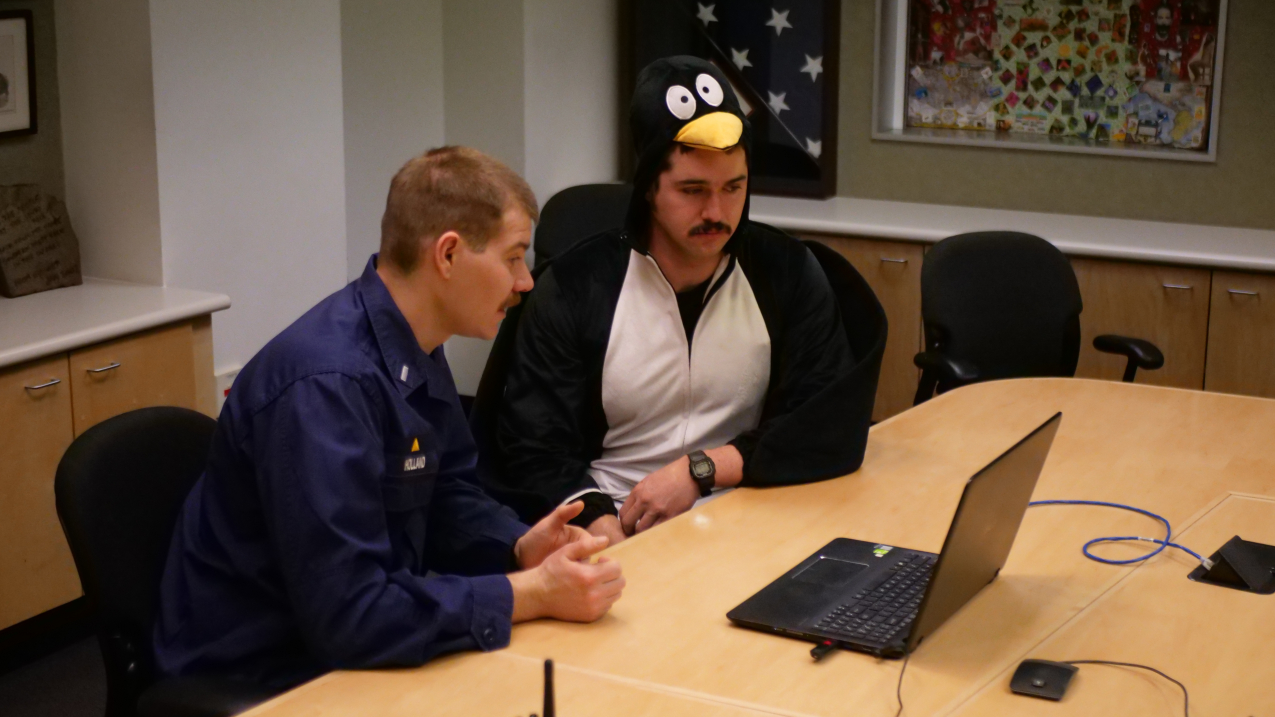 Two people, one in a NOAA Corps uniform and one in a penguin costume, sit in front of a laptop in a conference room.
