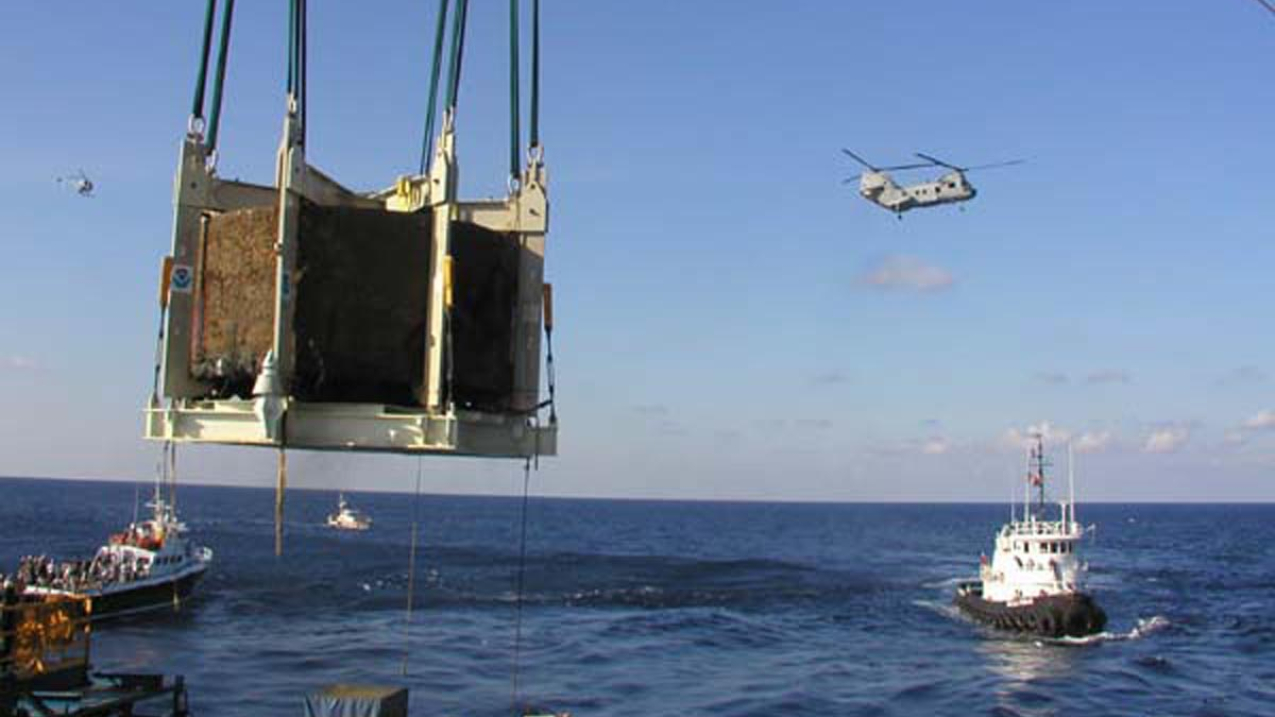 A large boom filled with a brown cargo box is hanging over the ocean while two tug boats wait in the background. A helicopter can also be seen in the sky. 