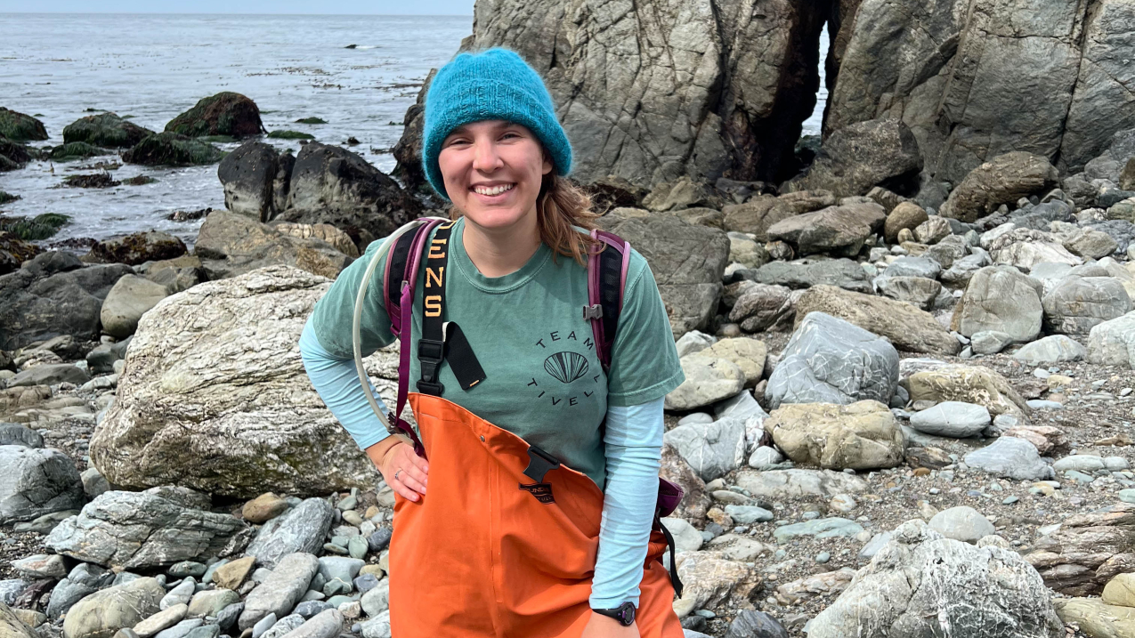 Audrey smiles with her hand on her hip, wearing a pair of bright orange waders, a winter hat, and long sleeves. She is standing in a rocky intertidal area characteristic of the Pacific coast. The water looks choppy the sky is overcast.